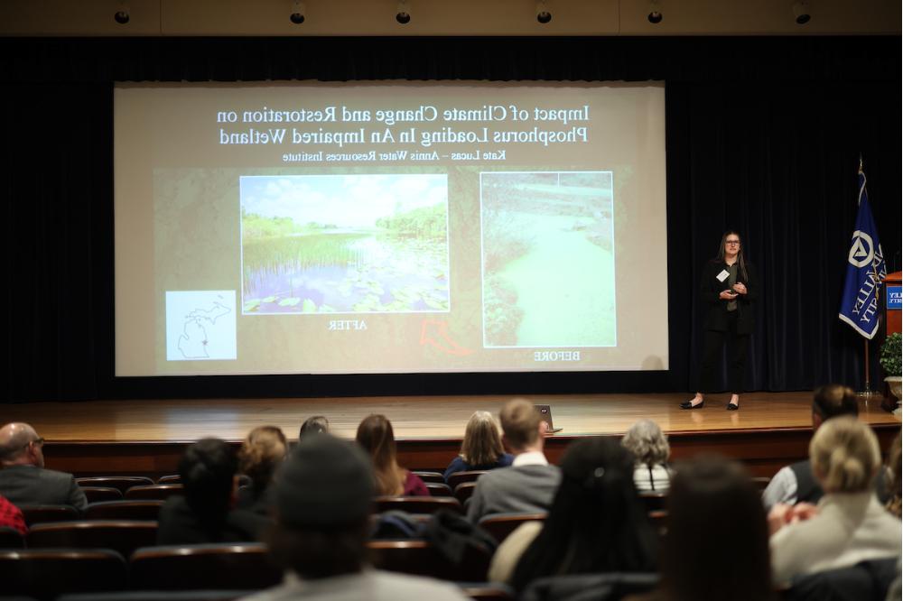 Kate Lucas  stands on stage next to a projection screen with her research slide on it.