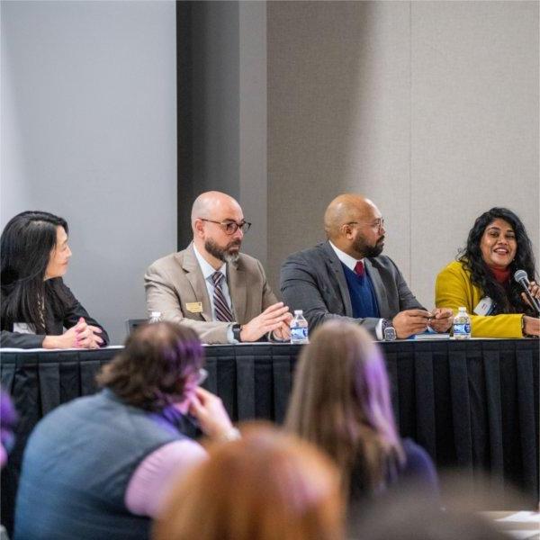 four people are panelists at a table, woman on far left with microphone