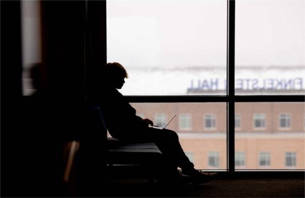  A college student is silhouetted against a window while working on a laptop. 