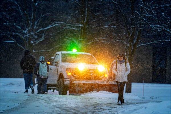   Students in winter clothing walk past a vehicle clearing snow from a snowy college campus. 