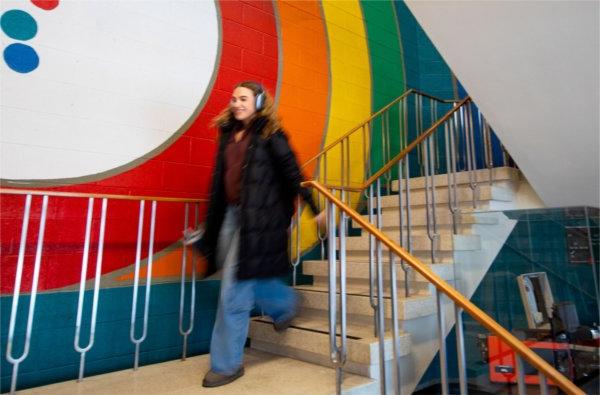  A smiling student walks past a colorful rainbow mural inside an academic building. 