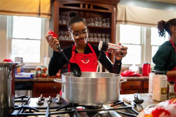  A smiling student wearing a red apron sprinkles spices into a pot. 