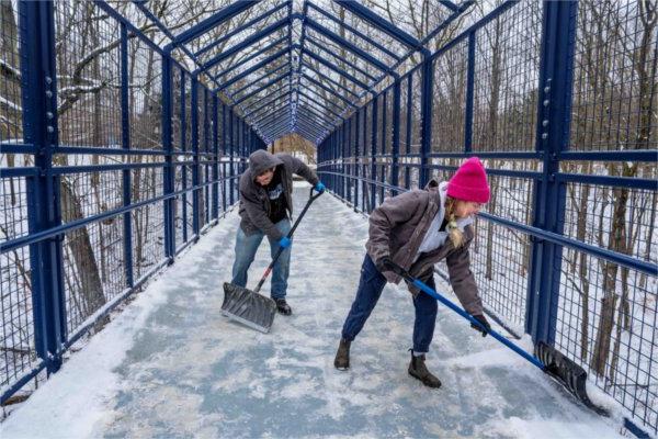  Two people use shovels to clear snow on a blue bridge. 