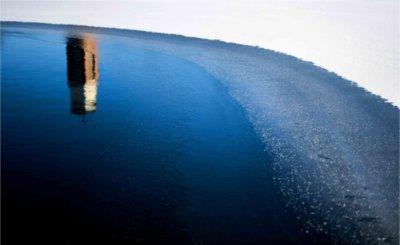 A reflection of a carillon tower is seen in blue water near frozen ice.