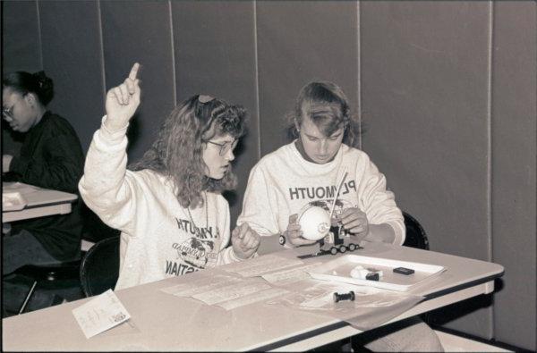 Two people wearing shirts that say "Plymouth Christian" sit at a table. One person is raising a hand.