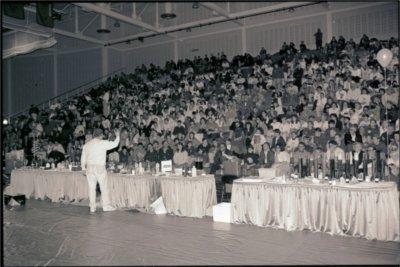 A person stands before a crowd seated in bleachers. Tables covered with cloths contain trophies and other items.