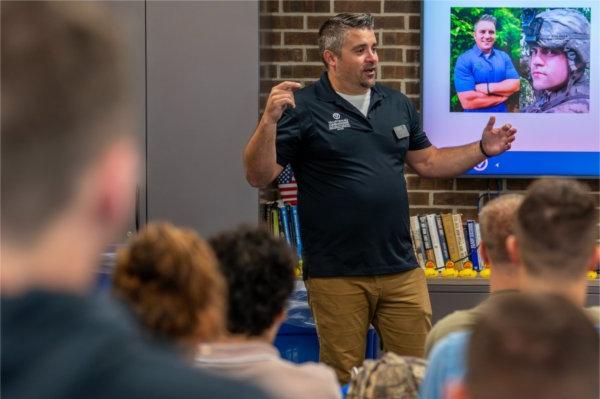 A person gestures while speaking before a group. There are pictues of the person on a screen in the background.