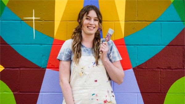 Rhiannan Sibbald wears paint-stained overalls and holds a paint brush. She stands in front of a colorfully-painted brick wall.