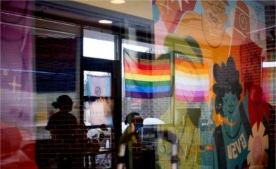 Through a piece of plexiglass, the reflection of two people and a pride flag can be seen, overlapping with the mural being installed on the LGBT Center&rsquo;s wall.