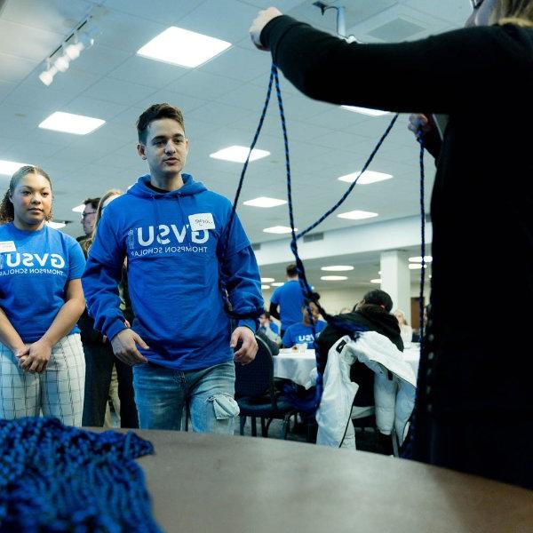 students in blue t-shirts with GVSU Thompson Scholars stand in line to receive graduation cords, which are blue