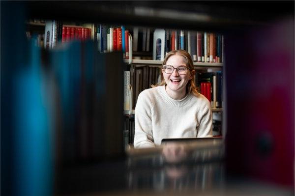 A person, seen through an opening of books on the shelves at a library, looks out of frame while laughing. 