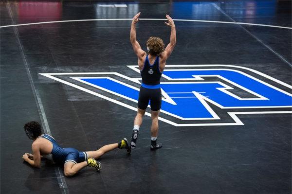 A person stands on the mat with his hands in the air in victory after a wrestling match while his opponent is still on the ground. 