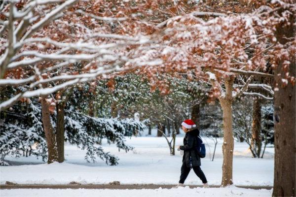 A person walks on a sidewalk in snowy surroundings wearing a Santa hat.  