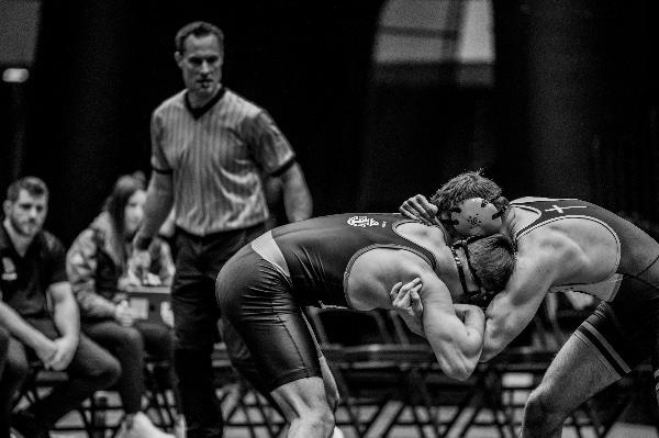 Two wrestlers are crouched with heads and arms entangled while a referee watches from behind them. 