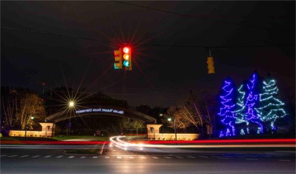 Holiday lights decorate the entrance to campus as traffic lights are blurred with a long camera exposure.