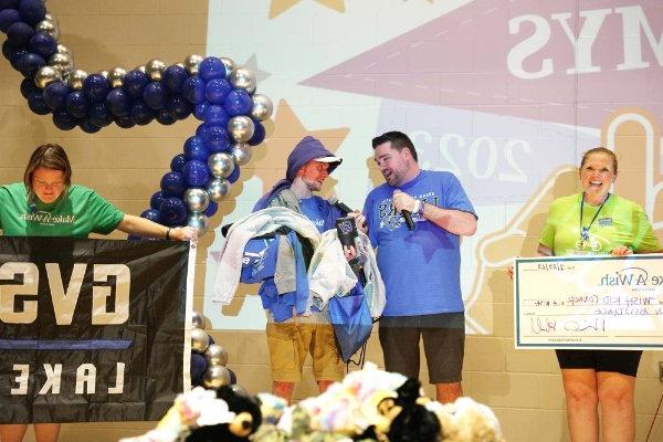 Connor Pung stands on stage holding a pile of GVSU clothing. Michael Hull stands next to him with a microphone, and two volunteers hold up a giant check and a GVSU Lakers flag.