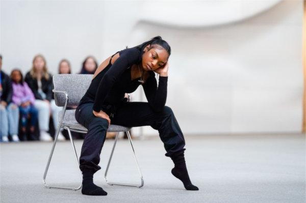   A dance and theatre performer wears all black and sits in a chair as they combine their talents during a performance.