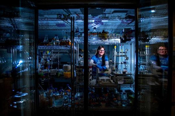 A college student wearing safety glasses poses for a portrait in a cold room of a biochemistry lab. 