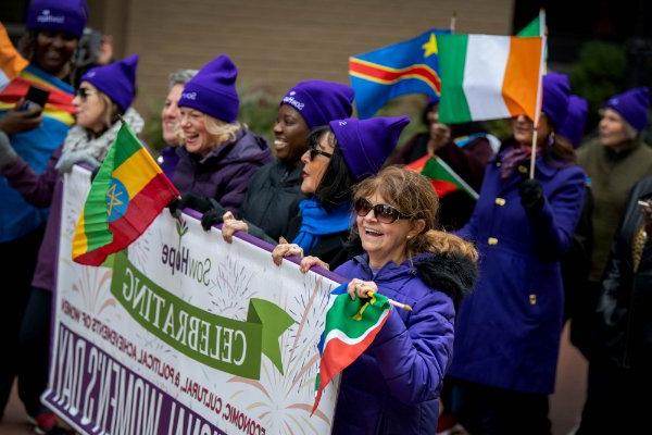 A group of women march wearing purple hats holding a banner and colorful international flags during a march through town.