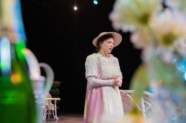 A actor wearing a white and pink dress and straw hat performs on stage near white wicker furniture during a play.