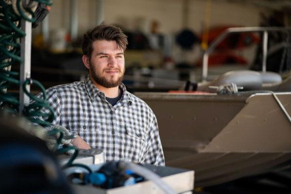 A student stands in a large garage structure filled with boats and boating gear for research. He looks out toward the light with a serious expression. 