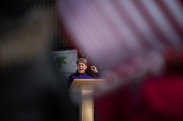 A speaker framed by the American Flag stands behind a lectern and speaks to a crowd.