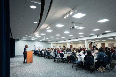 Kyra Harris Bolden speaks from a podium to an audience in a room with tables covered in white tablecloths.