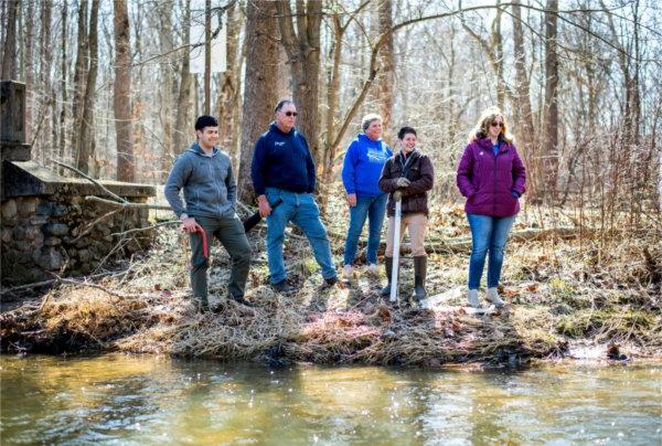 Five people standing along a creek bank smile while looking into the creek.