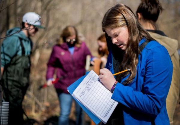 A person uses a pencil to write on a piece of paper in a folder while conducting research at a stream.