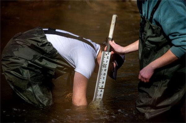 Two people standing in a stream place a ruler in the bottom of the stream.