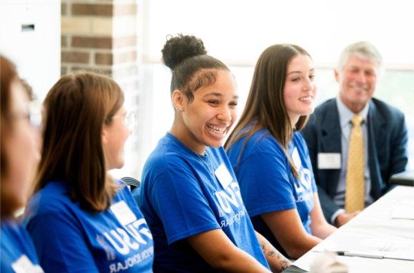 row of students in blue t-shirts, man in suit at end of table