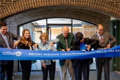 people stand behind a blue ribbon, Bob and Ellen Thompson Student Success Center, with scissors in hand to cut the ribbon