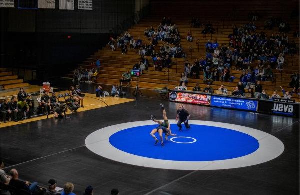 One wrestler on a mat flips the other as a referee and spectators look on.