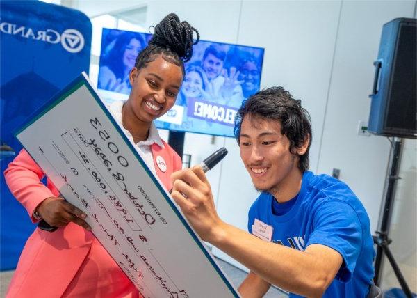 One person uses a marker to sign a large cardboard check held by another person. Both are smiling.