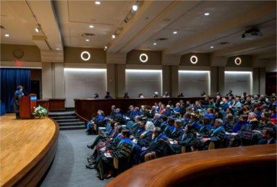 Samhita Rhodes, professor of engineering, speaks on stage in academic regalia to an audience at faculty convocation in Loosemore Auditorium