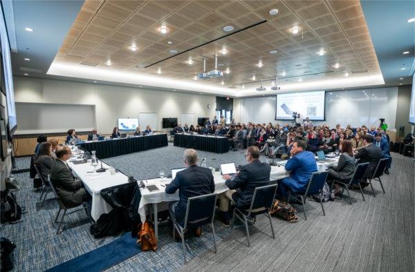 People sit at tables set up in a semi circle before a seated audience in a meeting room.