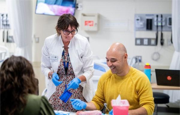 Henry Peña seated laughs with a professor in a white coat during a suturing lab