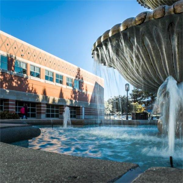 A water fountain sprays water on a sunny day. The Student Services building can be seen behind it.