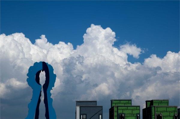  A blue art sculpture is seen in front of large puffy white clouds and a blue sky. 