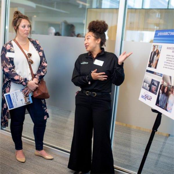 A person gestures toward a poster board while talking to others.