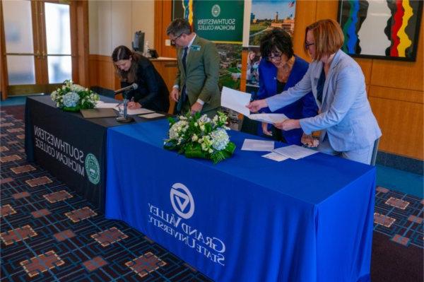 four people signing papers while standing behind two tables with draping for GVSU and SMC