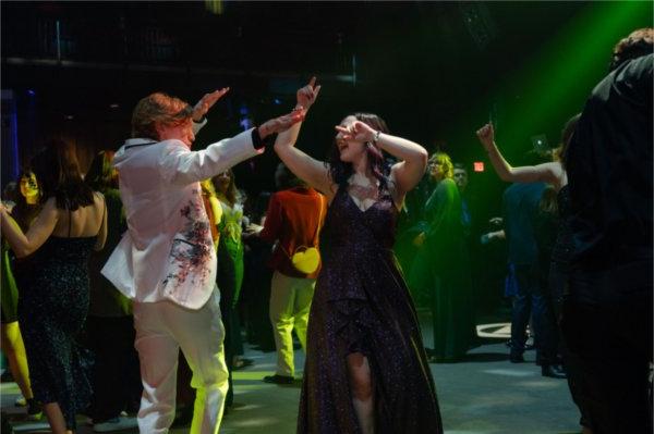 A young man in a beaded blazer and a young woman in a dark sparkly dress point at one another as they do disco dance moves on the dance floor.