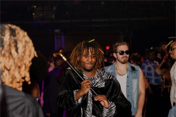 A young man in a black and zebra-striped 70s-style suit poses with a cane on the edge of the dance floor. He has two glow sticks stuck in his short dreads. 