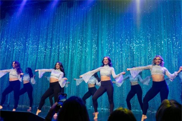 A group of dancers in white shirts with large sleeves perform in front of a sparkly blue curtain. 