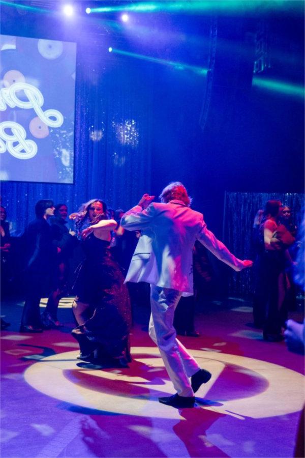 A young man in a white suit and young woman in a red dress do a lively dance in the middle of the dance floor. 