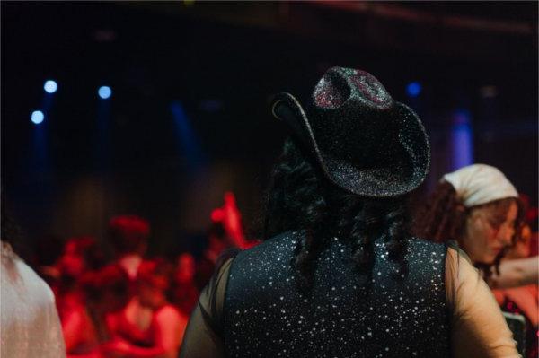 A person wears a black sparkly cowboy hat and black sparkly vest.