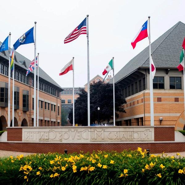 Several flags on flagpoles fly outside of a building. A cement sign says "Richard M. DeVos Center."