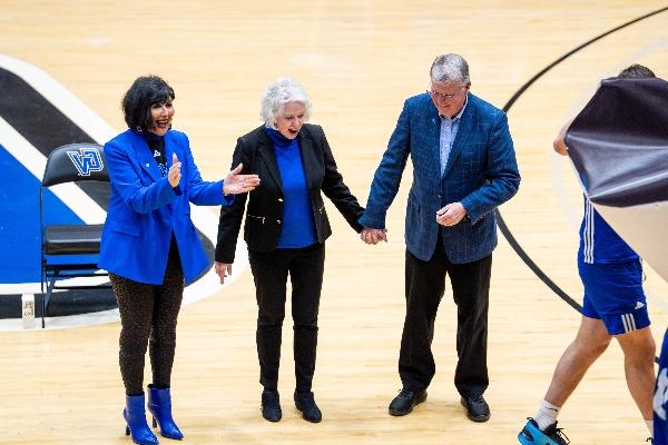  A university president wearing blue claps as a basketball court named after a former university president, pictured holding hands with his wife, is unveiled. 