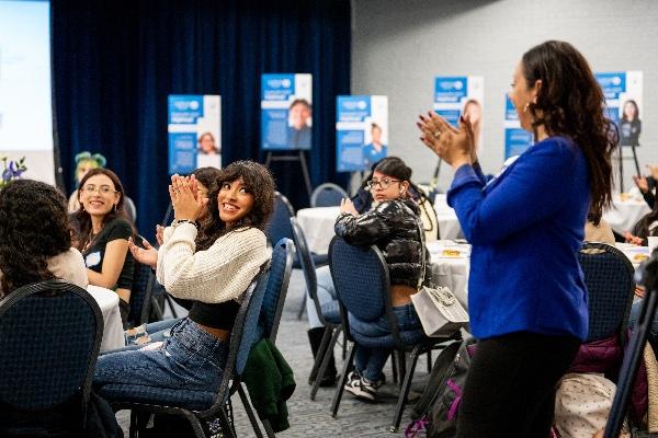  A room full of college students and participants clap and laugh together. 