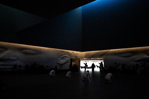 Dancers are silhouetted in an entrance to a room with face sculptures on the wall. 